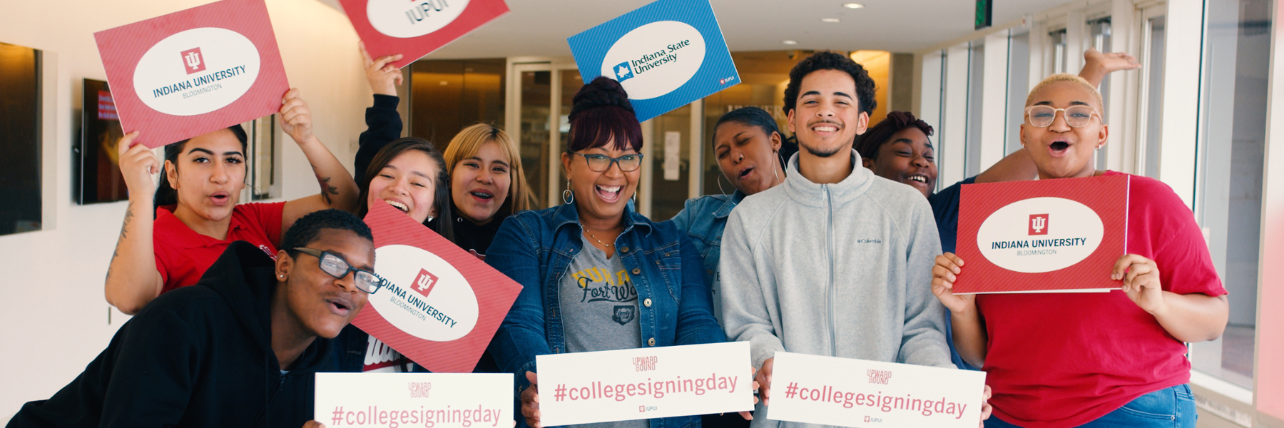Students posing at College Signing Day while holding signs from various universities.