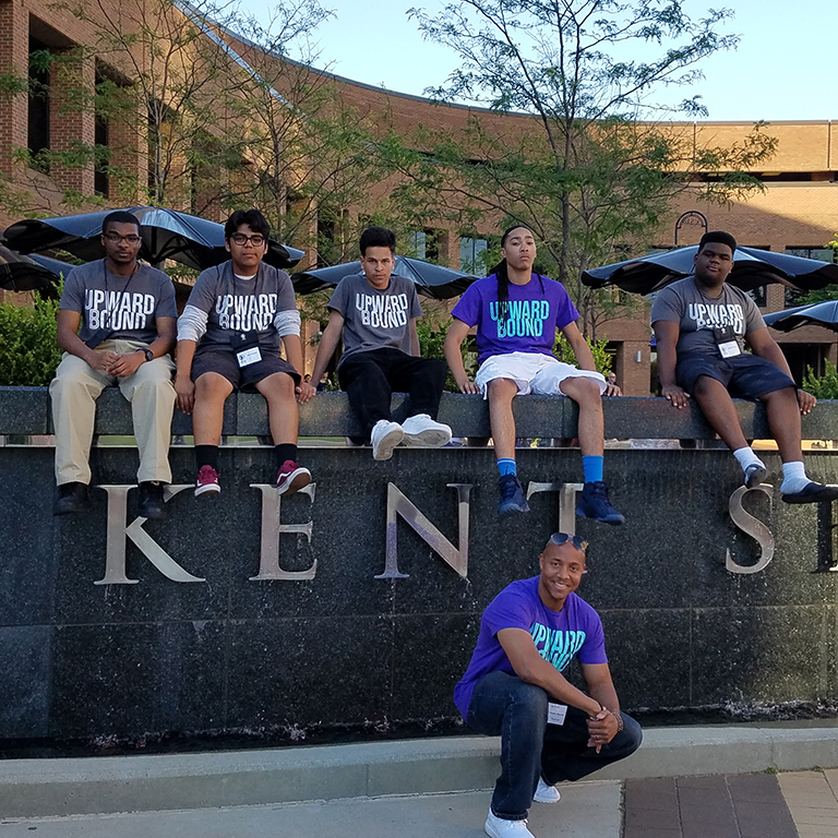 Upward Bound Men of Excellence participants sitting on the Kent State sign. 